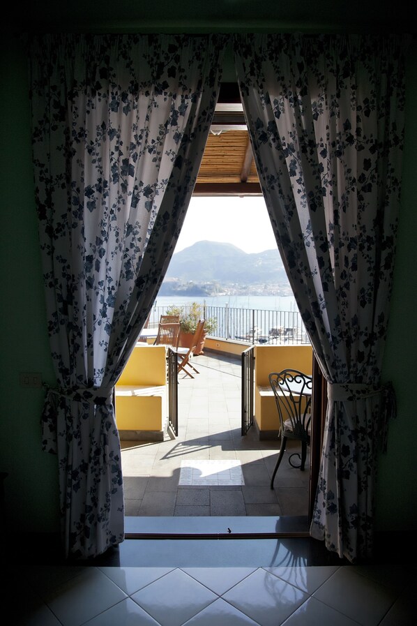 Vista a través de una ventana con cortinas florales, que da a un balcón con sillas amarillas y una mesa, con vista a una masa de agua y montañas.