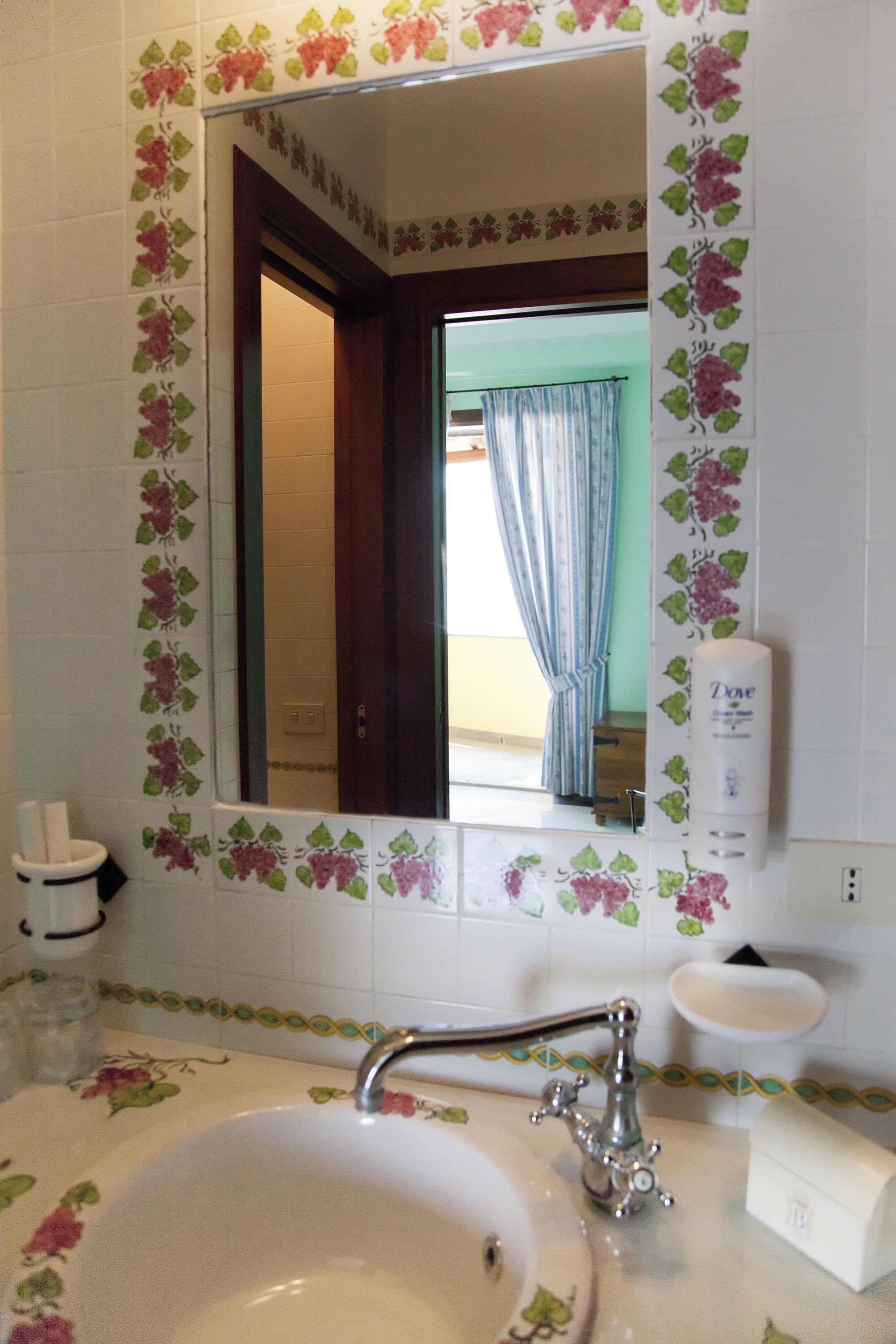 Une salle de bain avec une bordure de carrelage à motifs floraux, un lavabo avec un robinet, un miroir et une fenêtre avec des rideaux.