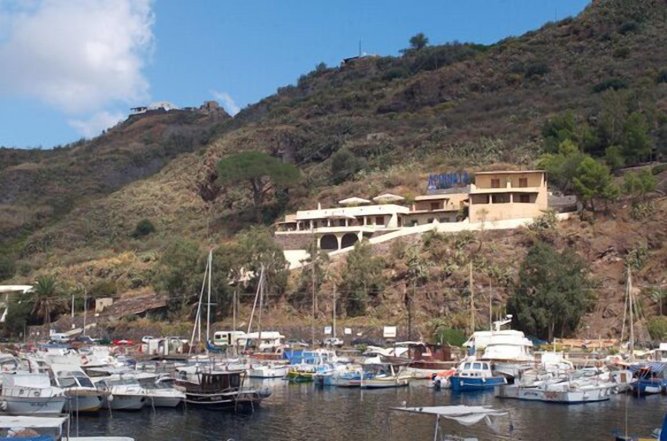 Un port de plaisance avec des bateaux amarrés, un bâtiment portant une enseigne, et un paysage vallonné en arrière-plan.