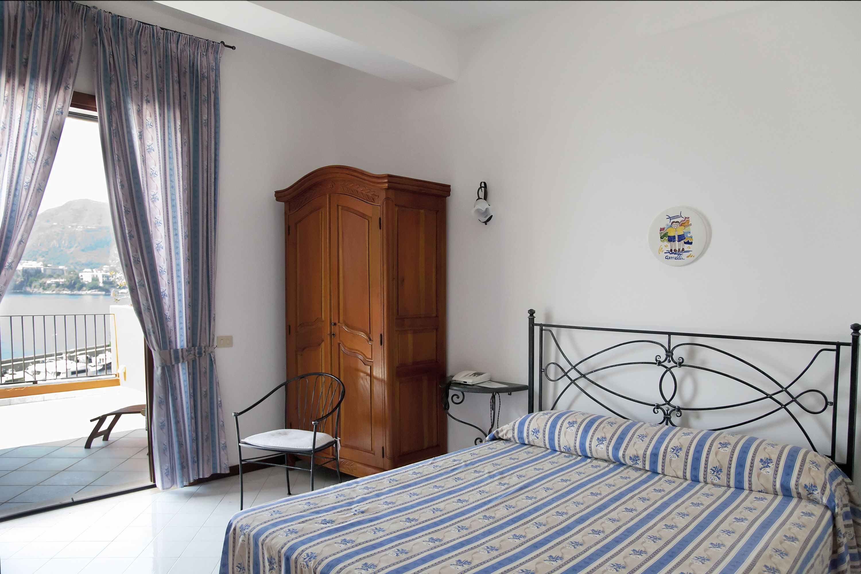A bedroom with a bed, a wooden wardrobe, a chair, a small table, and a balcony with a view of the sea.