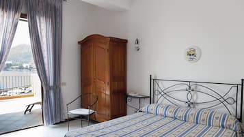 A bedroom with a bed, a wooden wardrobe, a chair, a small table, and a balcony with a view of the sea.