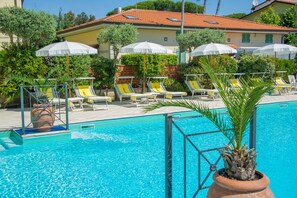 Zona de piscina con sillas de descanso y sombrillas, rodeada de vegetación y un edificio con techo de tejas.