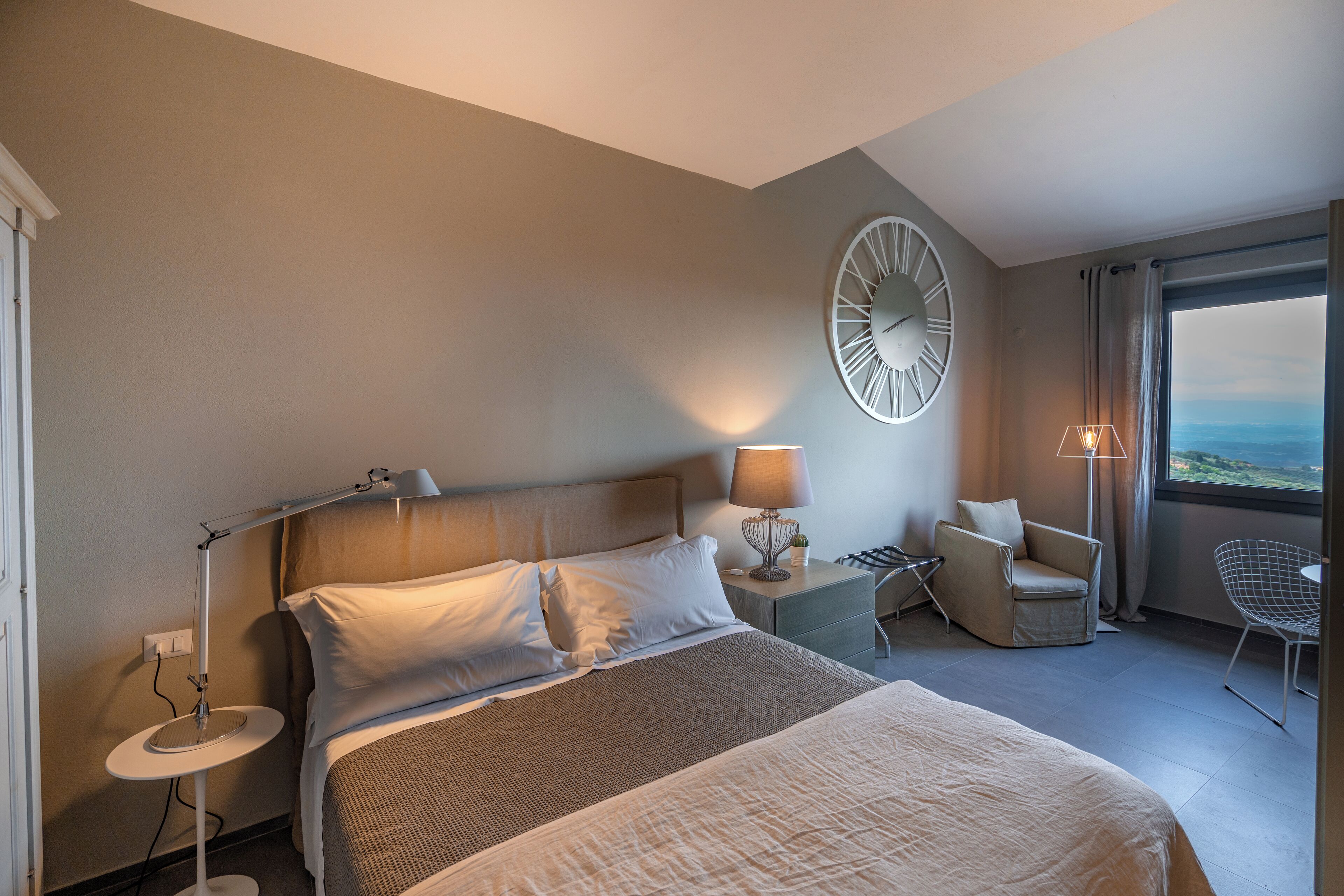 Une chambre d’hôtel avec un lit, des tables de chevet, une lampe, une chaise, une horloge et une vue sur la mer.