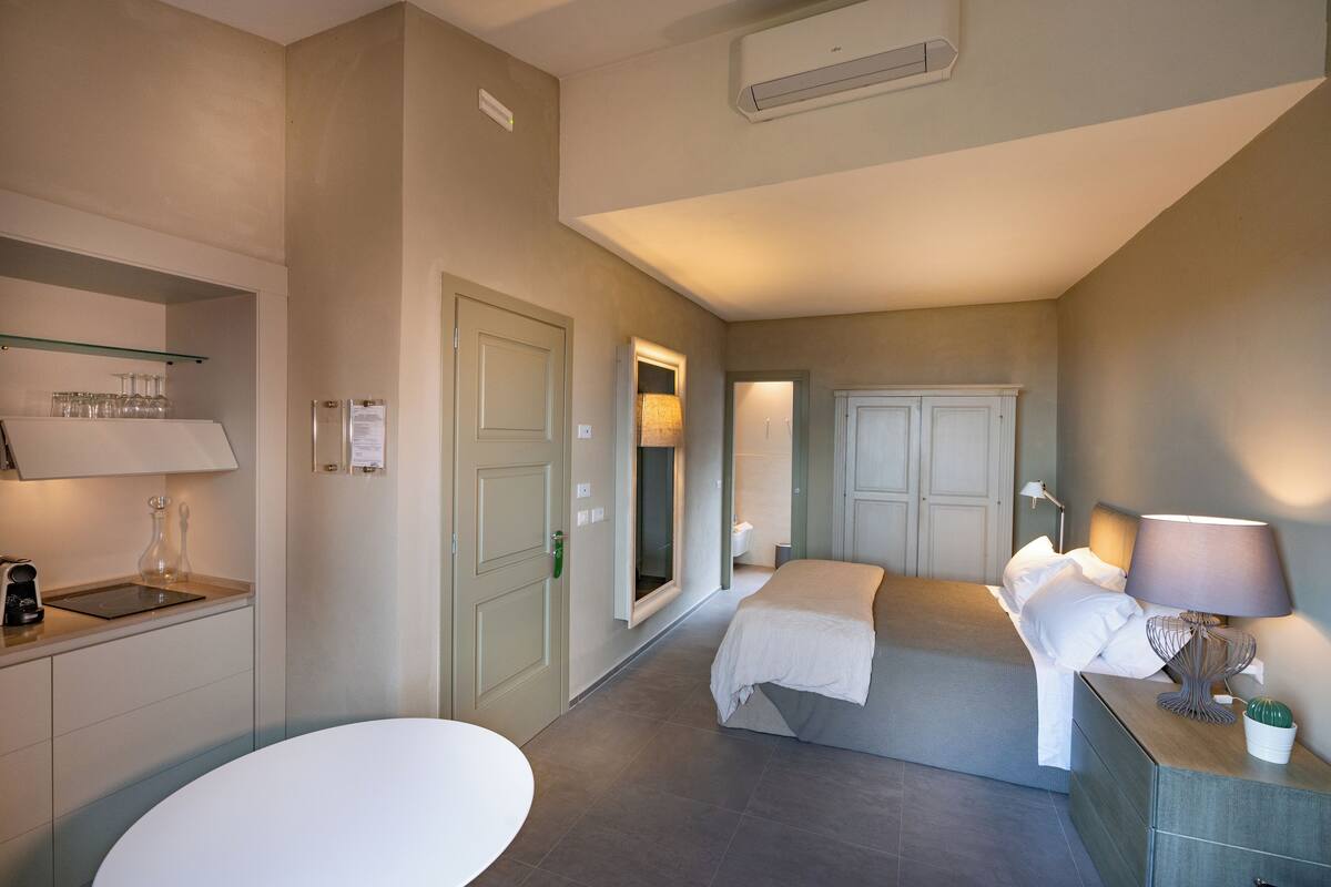 Ein Hotelzimmer mit Küchenzeile, einem Bett mit weißer Bettwäsche, einem Nachttisch mit Lampe und einer kleinen Topfpflanze.