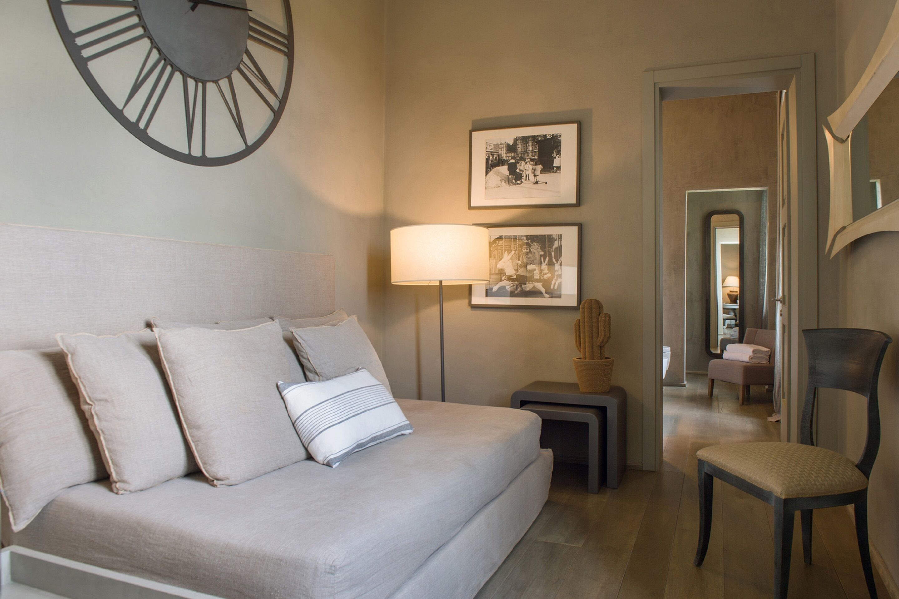 Una camera d'albergo con un letto, una sveglia a muro, quadri incorniciati, una lampada, un cactus e una sedia.