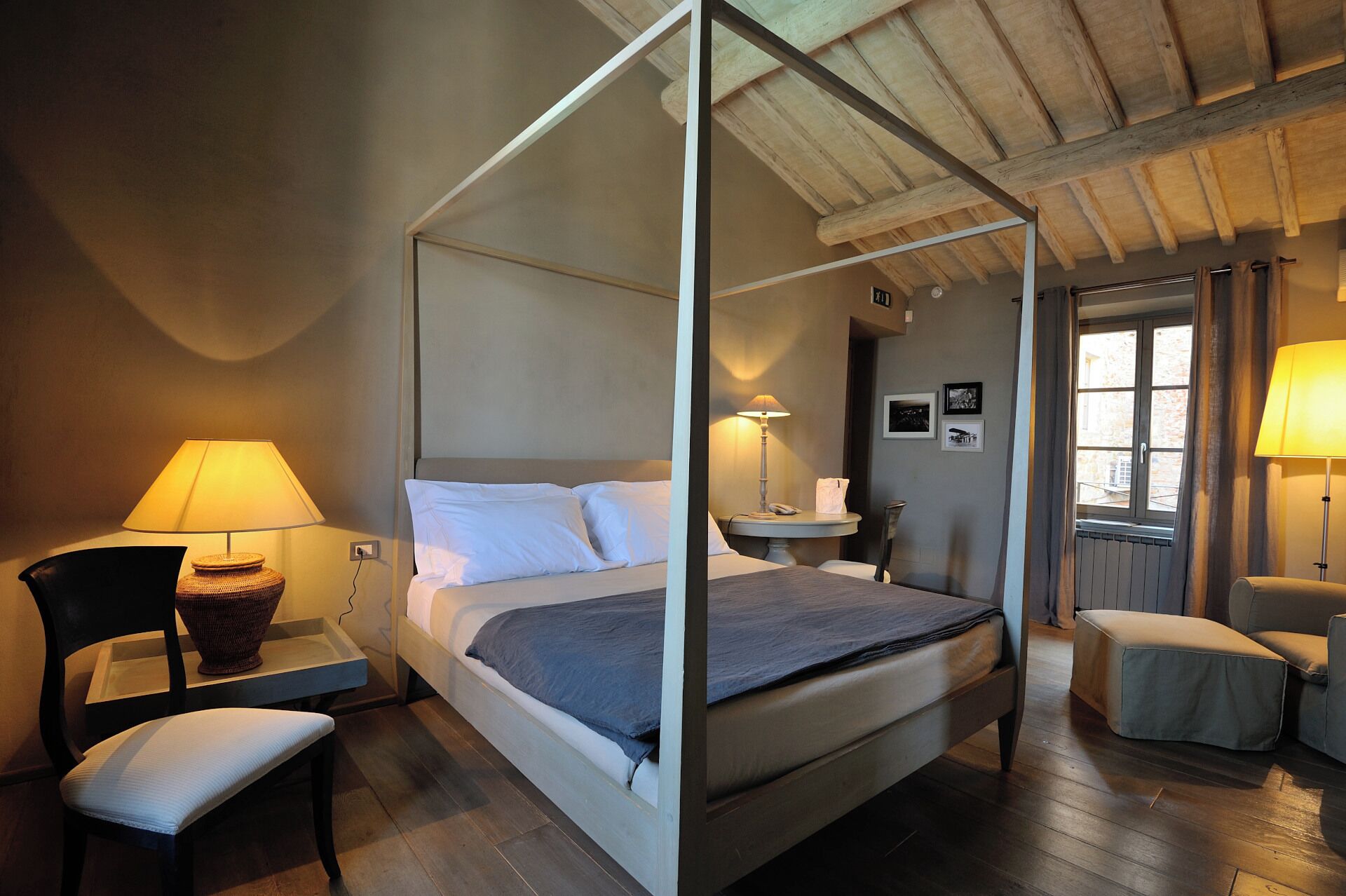 A four-poster bed with white linens, a bedside table with a lamp, a chair with a cushion, and a window with curtains.