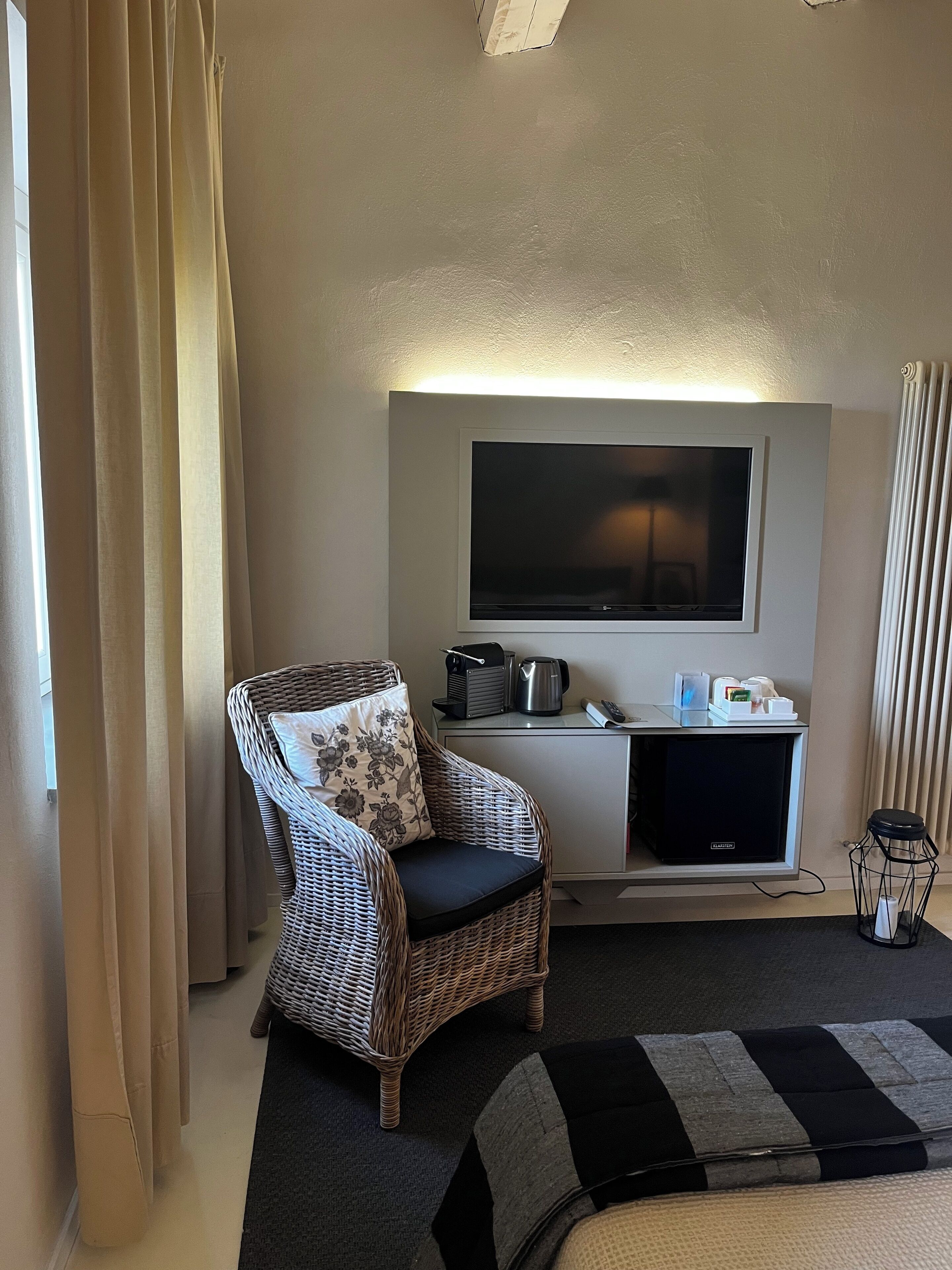 A hotel room with a wicker armchair, a flat-screen TV, a minibar, and a radiator.