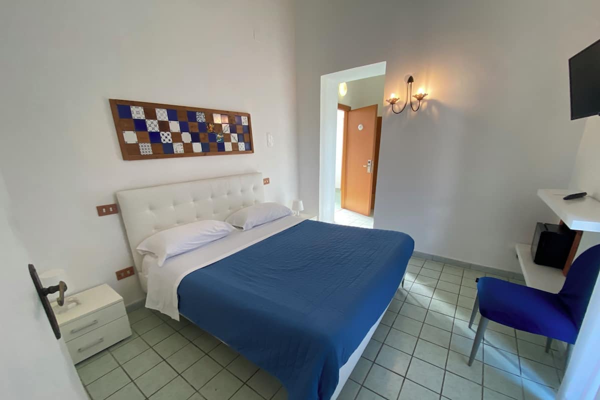 A bedroom with a bed, a desk, a chair, a wall-mounted artwork, and a door leading to another room.