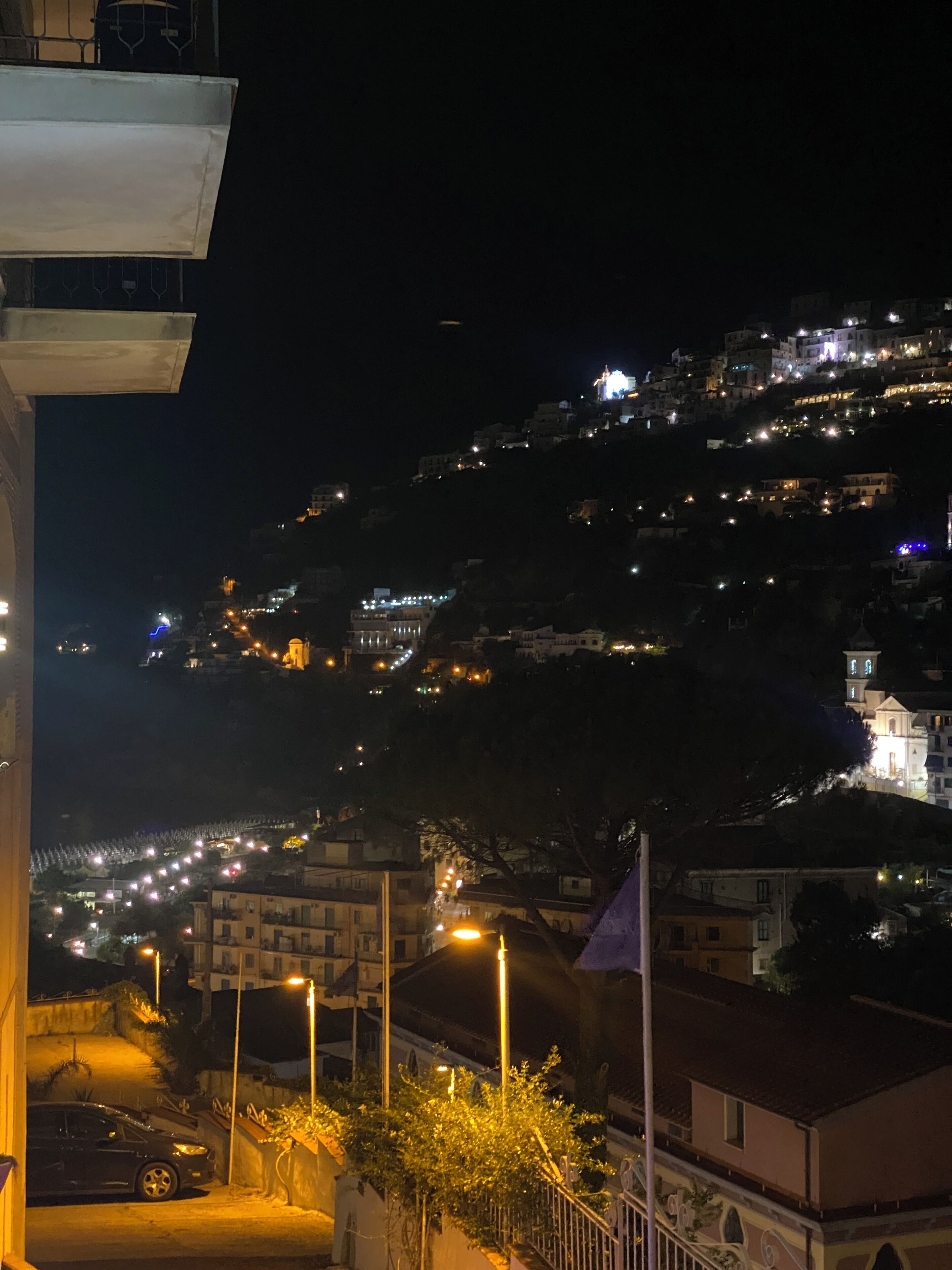 Vista nocturna de un pueblo en la ladera de una colina, con edificios iluminados por farolas y luz ambiental.