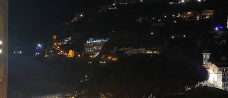Vista nocturna de un pueblo en la ladera de una colina, con edificios iluminados por farolas y luz ambiental.
