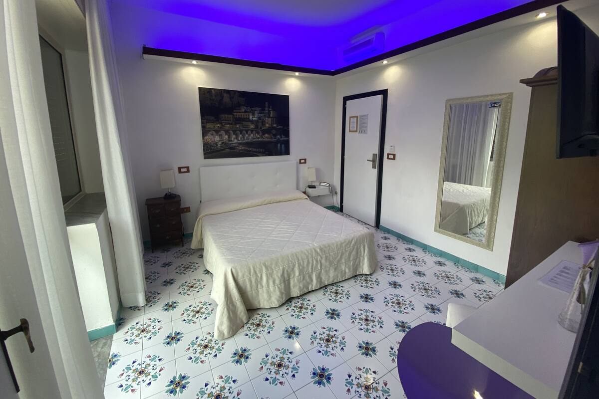A hotel room with a bed, a desk, a mirror, a ceiling fan, and a painting on the wall.