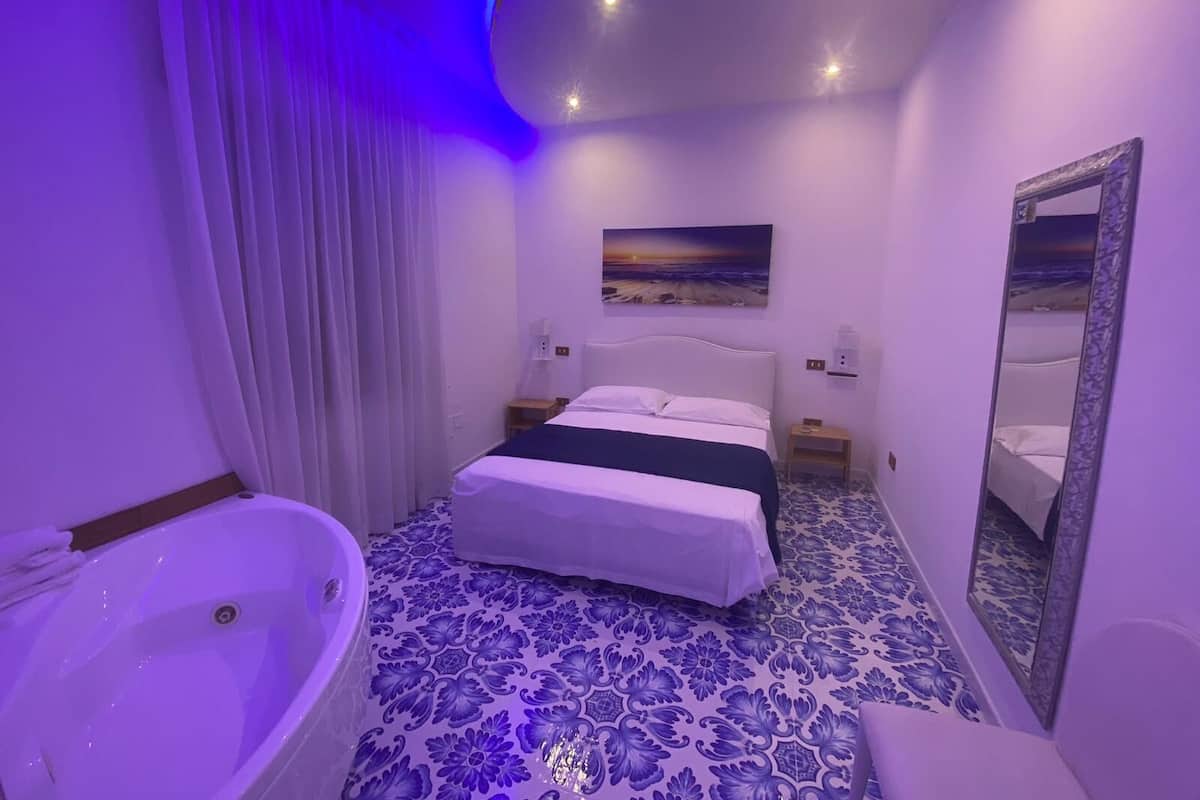 A bedroom with a bed, a nightstand, a wall-mounted mirror, a painting, and a bathtub.