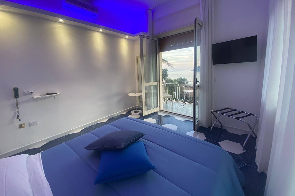 A modern hotel room with a bed, a TV, a balcony with a chair, and a view of the sea.