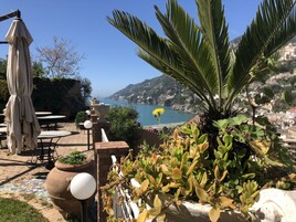 An outdoor seating area with tables and chairs, a potted plant, and a view of the sea and hills.