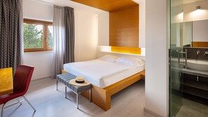 Habitación de hotel moderna con cama, una mesita con un sombrero, una silla roja, ventana con cortinas y baño con cabina de ducha de vidrio.