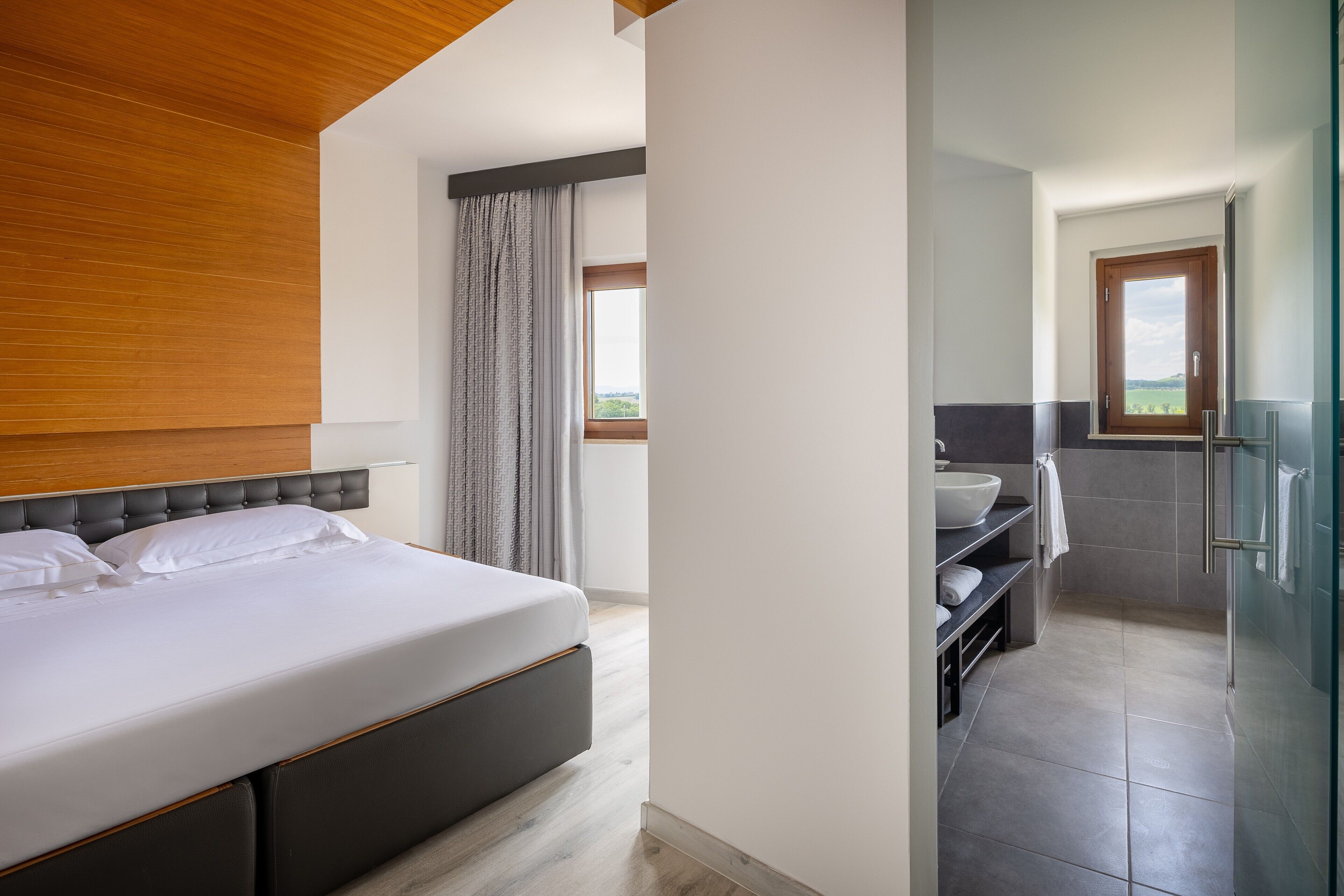 Ein modernes Schlafzimmer mit einem großen Bett, ein Badezimmer mit Waschbecken und Spiegel sowie ein Blick ins Freie.