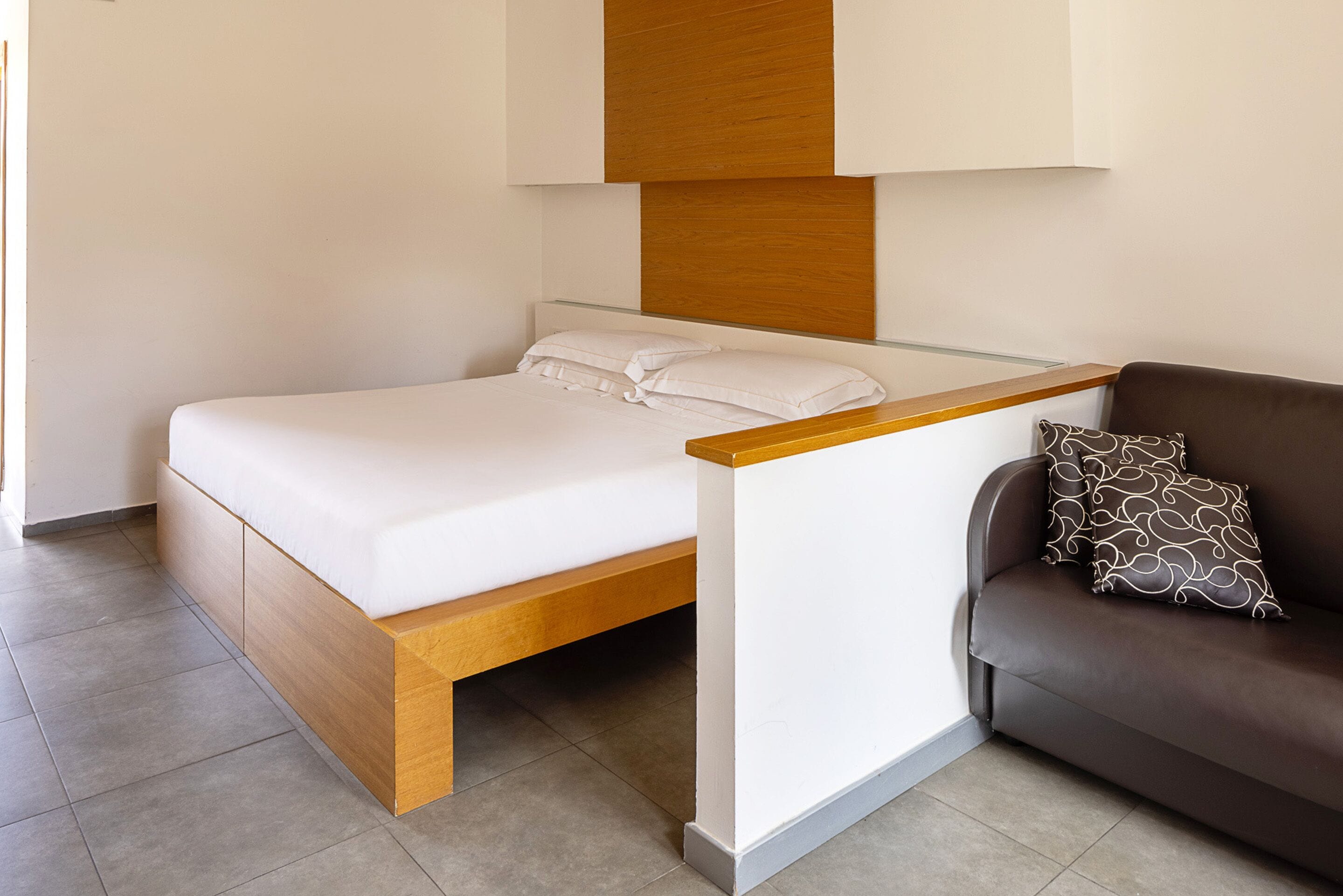 A modern bedroom with a wooden bed frame, white bedding, a glass headboard, and a brown sofa with patterned cushions.