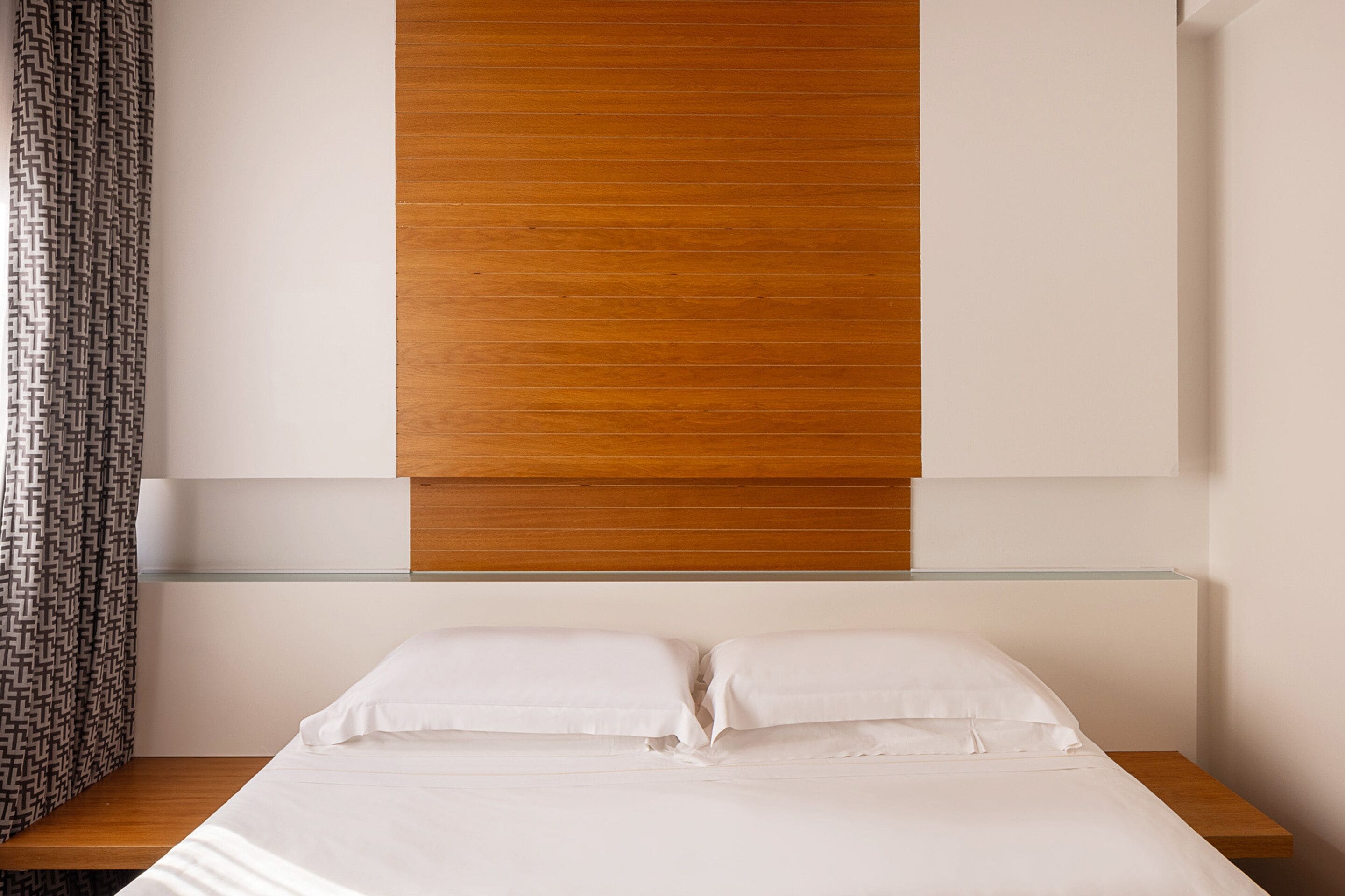 Een moderne slaapkamer met een houten hoofdbord, een glazen plank boven het bed en een kamerbreed gordijn met een patroon.