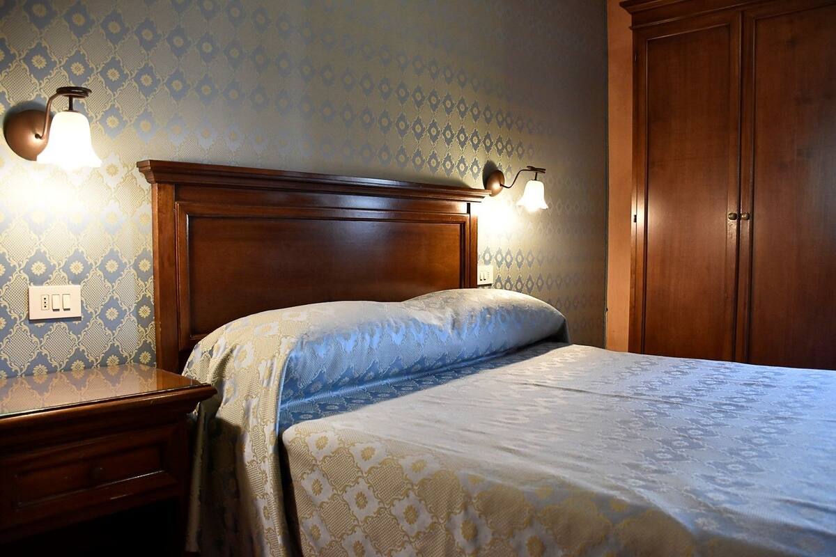 A bedroom with a wooden headboard, two bedside lamps, a bed with patterned bedding, and a wardrobe.