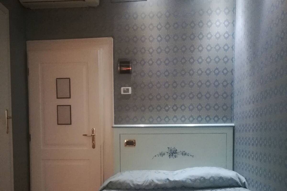 A bedroom with a patterned bedspread, a white door with a small window, and a wall-mounted air conditioner.