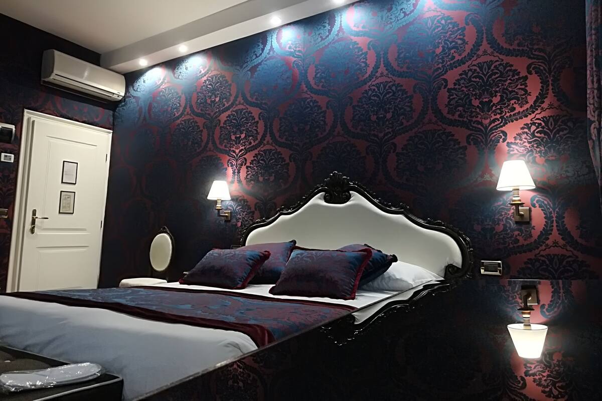 A bedroom with a dark wood bed, patterned wallpaper, two wall-mounted lamps, and an air conditioning unit.