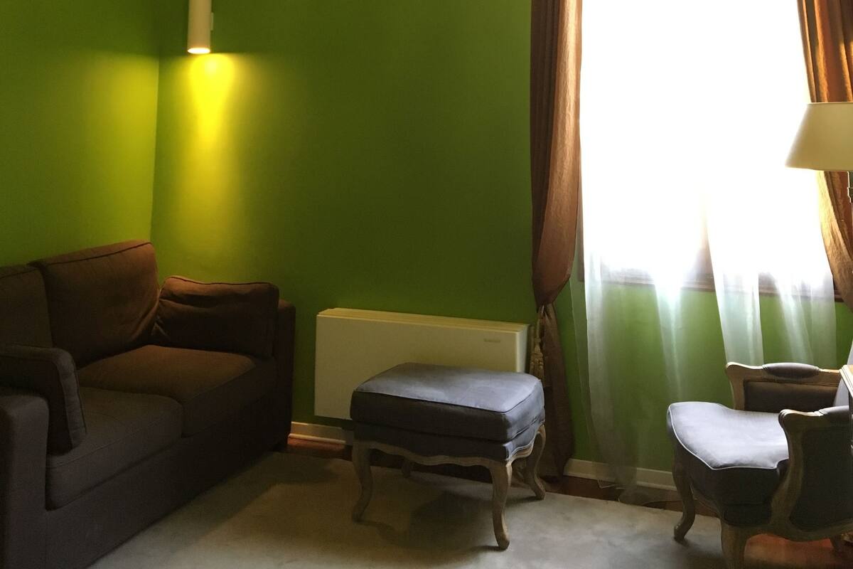 A room with a green wall, a chandelier, a radiator, a window with curtains, a sofa, an ottoman, and a chair.