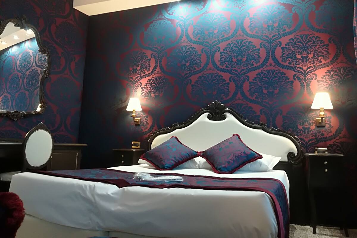 A bedroom with a large bed, ornate headboard, two bedside lamps, a round mirror, and patterned wallpaper.
