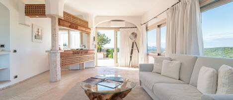 A spacious living room with a glass coffee table, a white sofa, and large windows offering a view of the landscape.