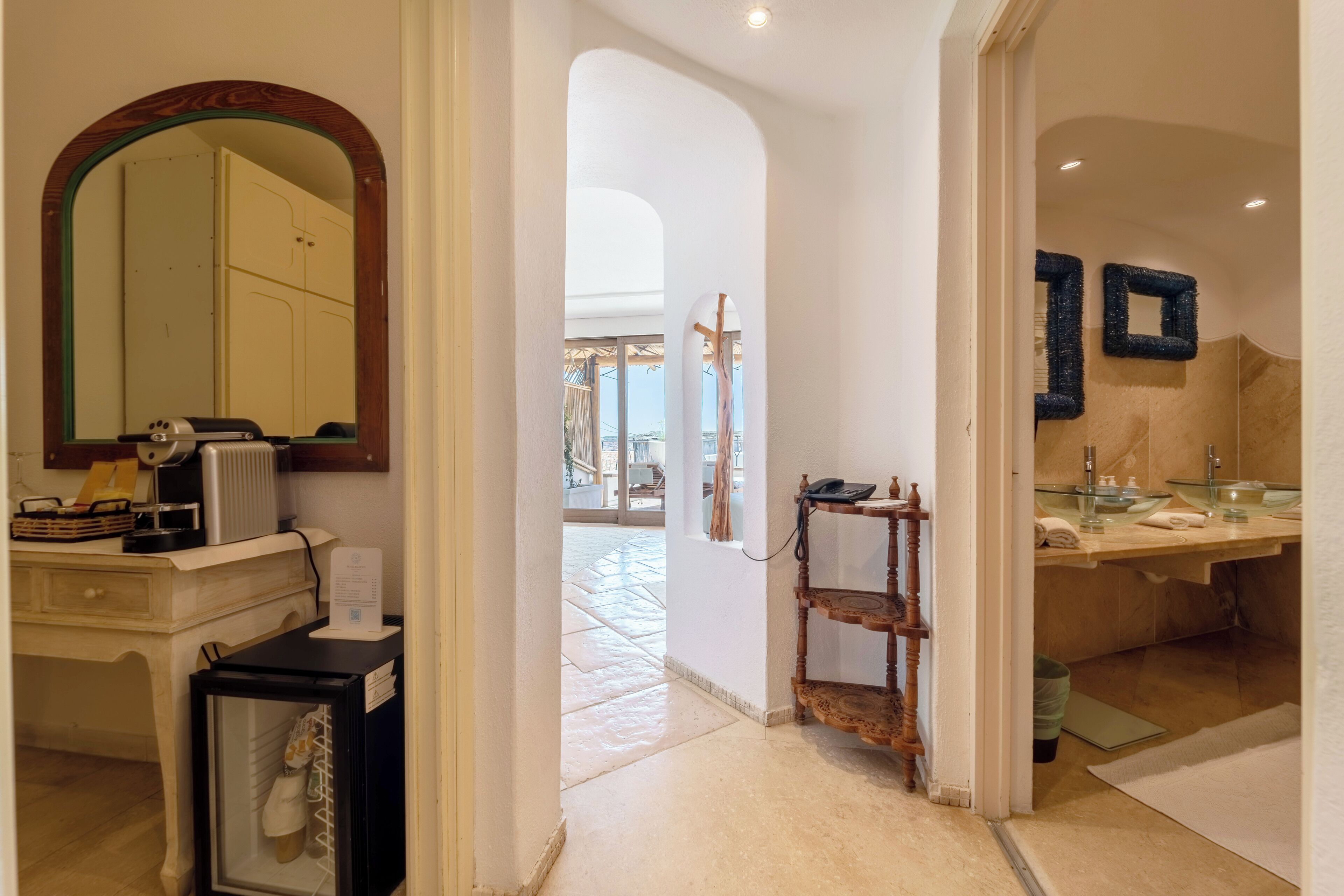 A hotel corridor with a mirror, a wooden shelf, and a bathroom with a sink and mirror.