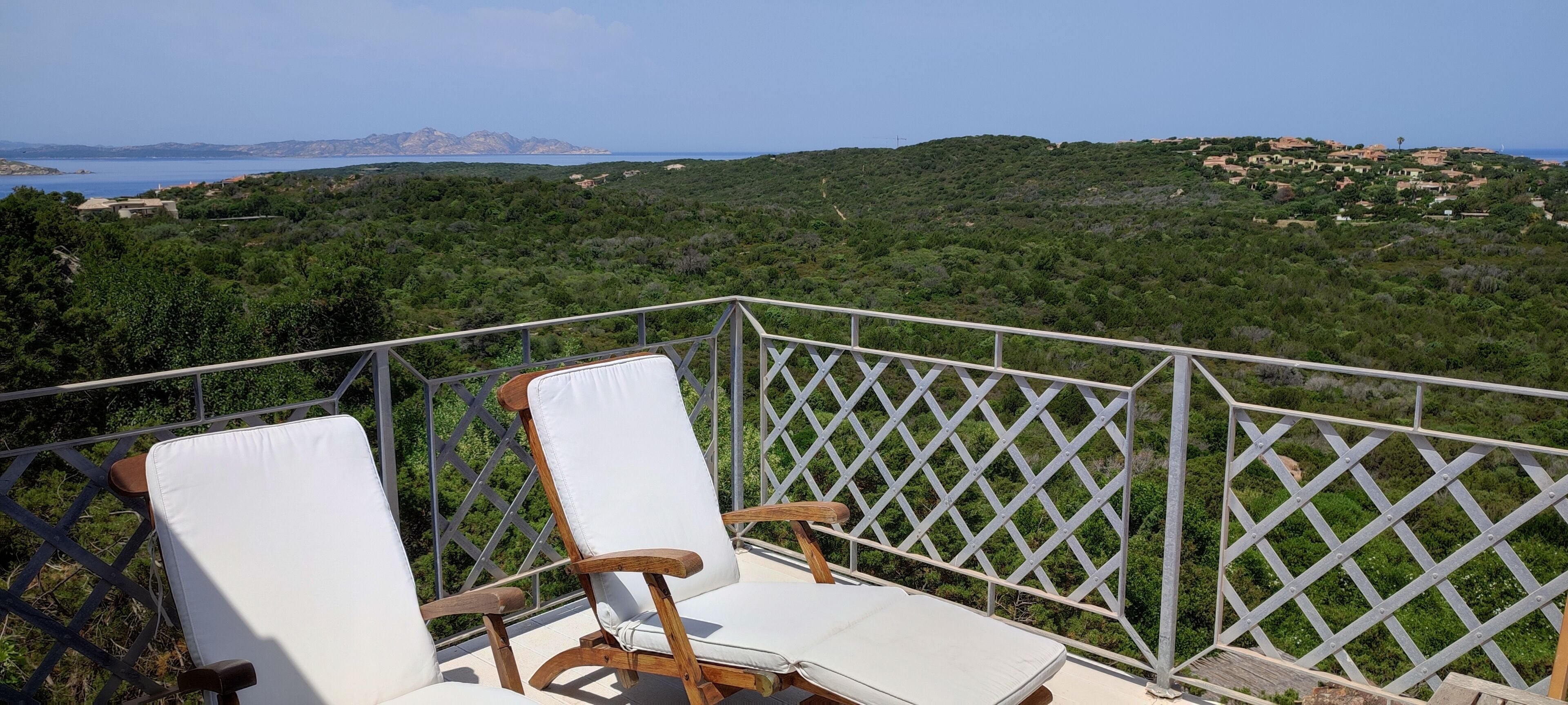 A balcony with two white cushioned chairs and a wooden table, overlooking a lush green landscape and a distant coastline.
