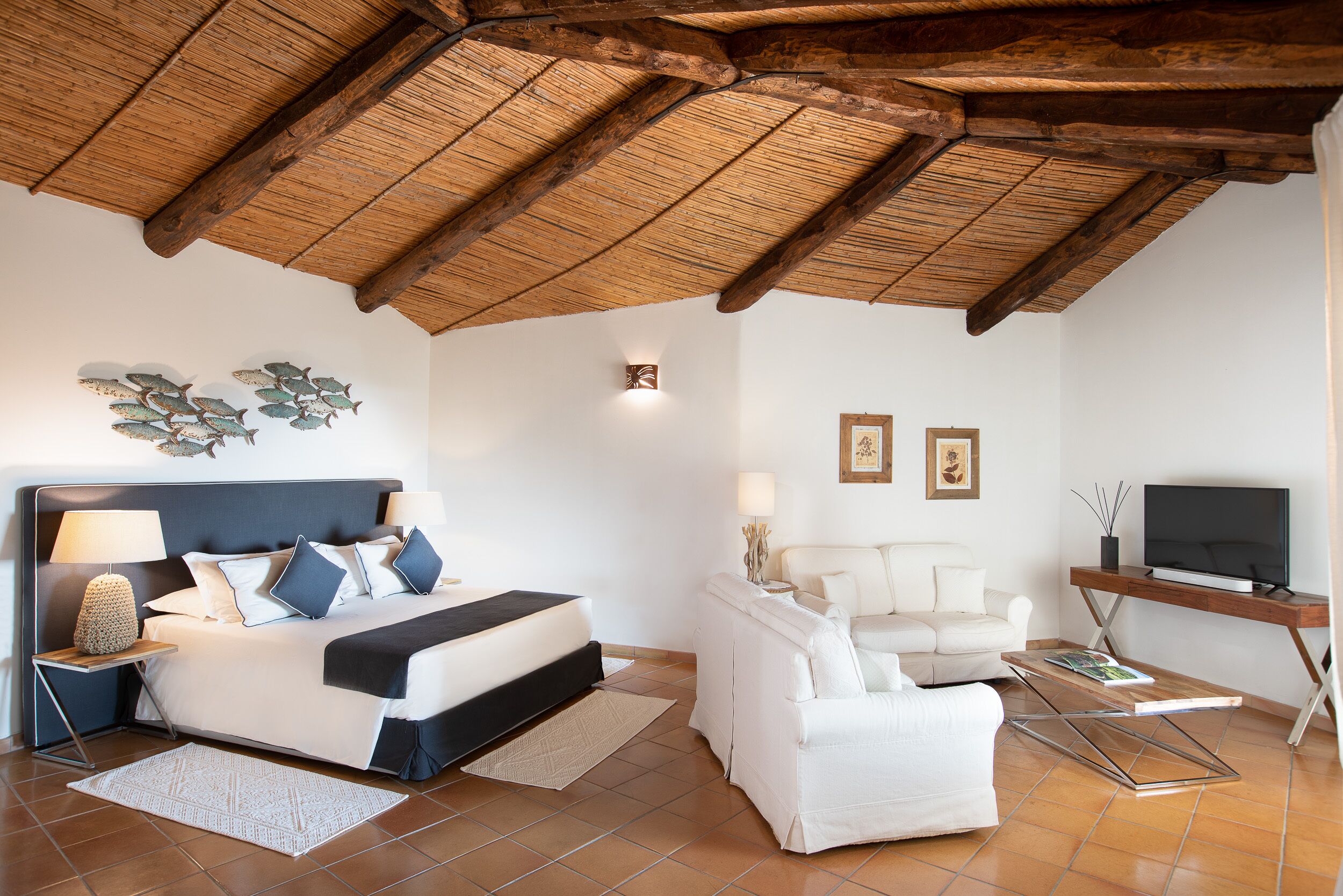 A modern bedroom with a wooden ceiling, a bed with white bedding, a white sofa, a TV on a wooden stand, and a small table with a lamp.