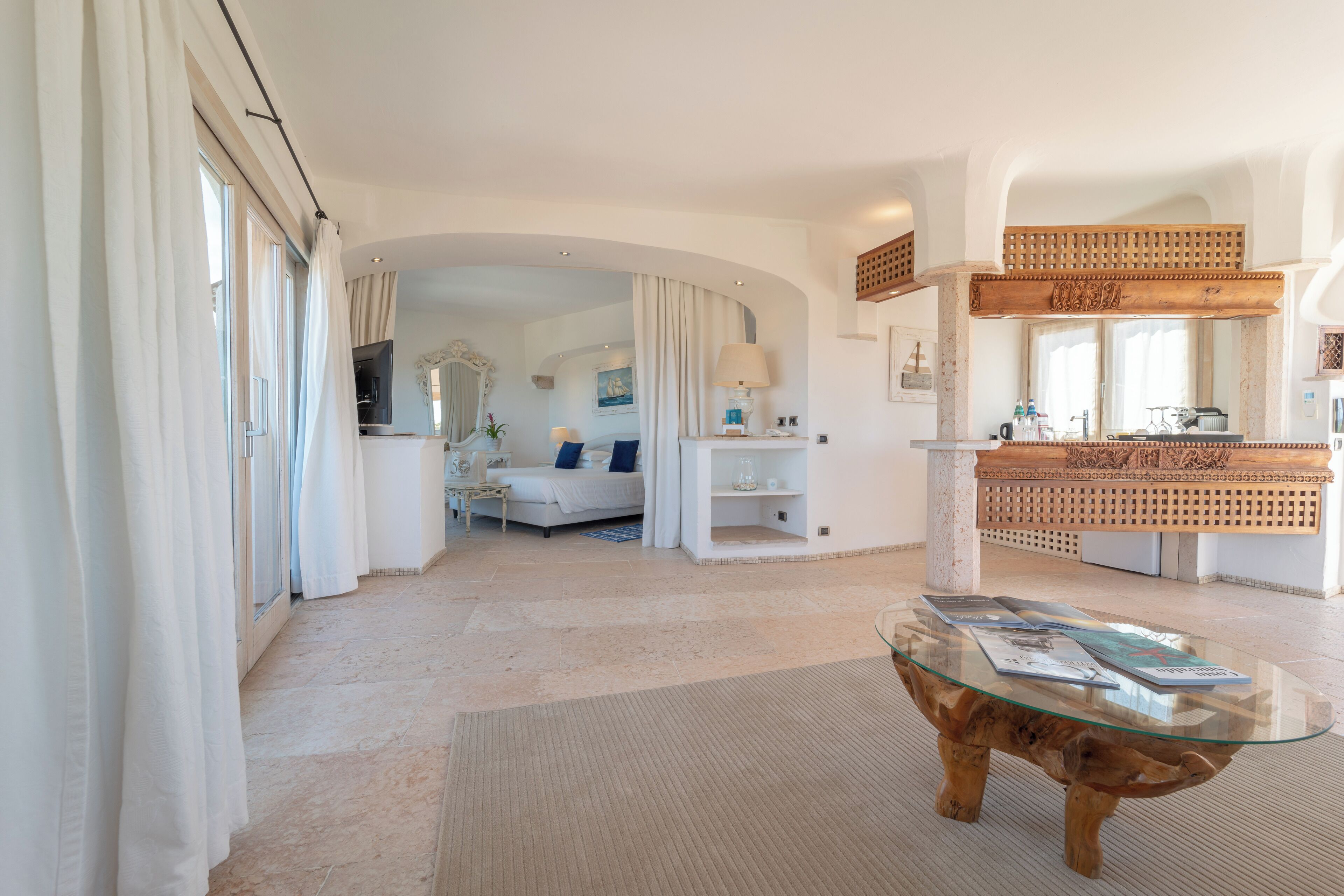 A spacious room with a glass coffee table, a bed with a blue accent pillow, and a bathroom with a wooden vanity and mirror.
