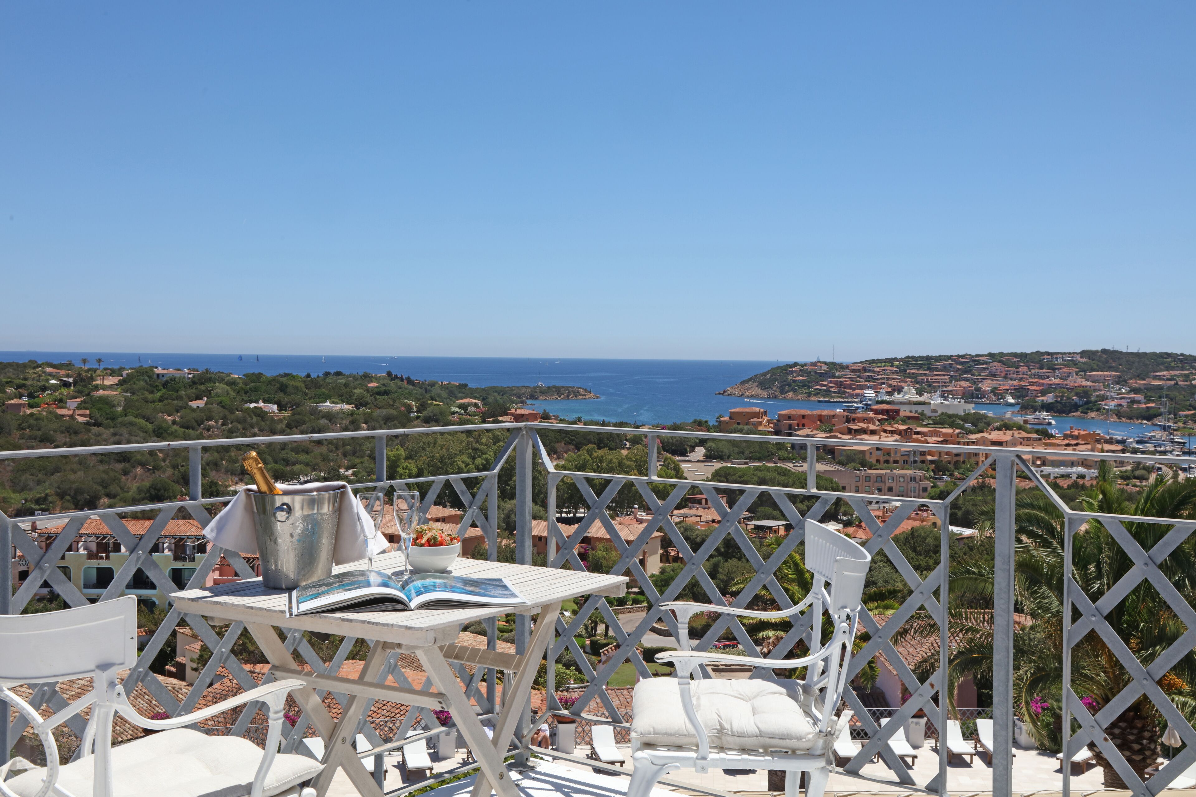 A terrace with white chairs and a table, overlooking a coastal town with buildings and the sea.