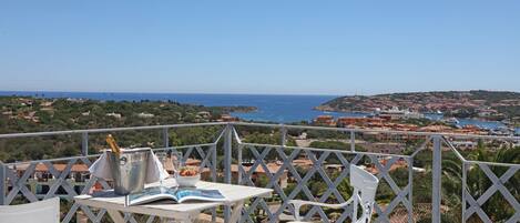 A terrace with white chairs and a table, overlooking a coastal town with buildings and the sea.