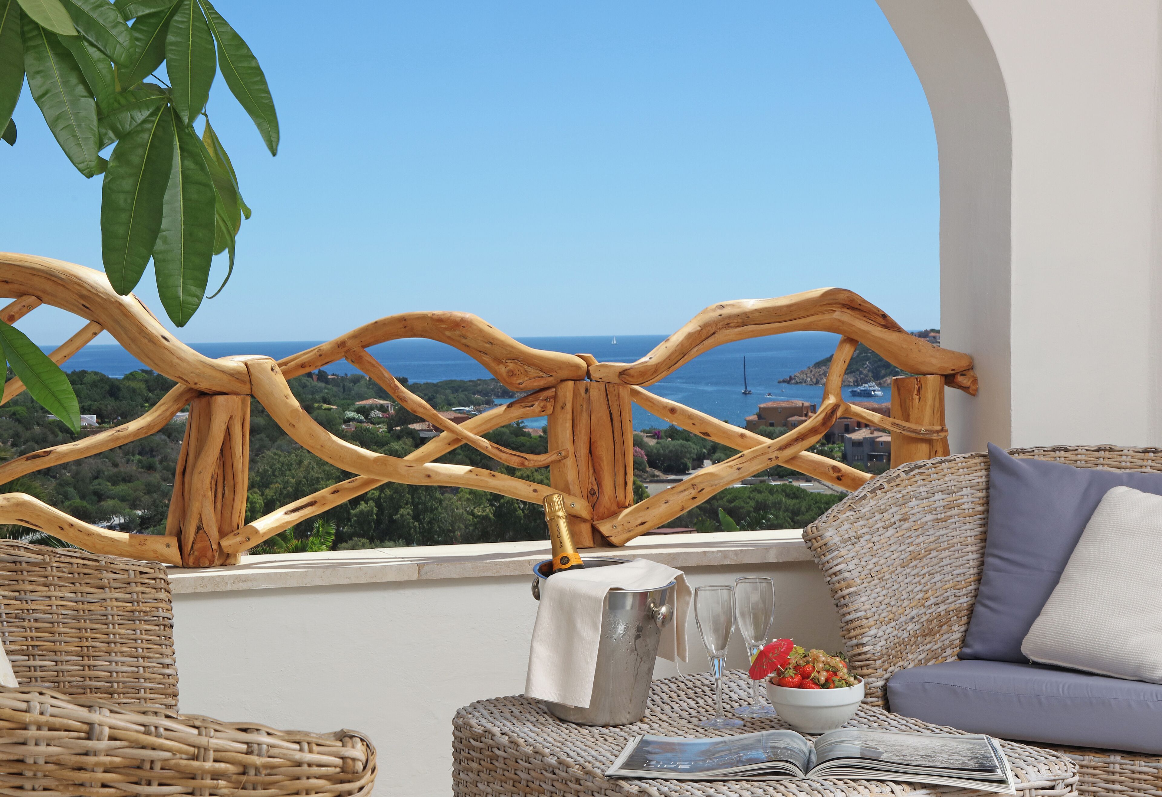 A wicker sofa with a cushion, a small table with a bottle, glasses, and a book, and a view of the sea through a wooden railing.