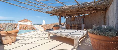 Un espacio al aire libre iluminado por el sol con piscina, macetas de terracota y una estructura de pérgola.