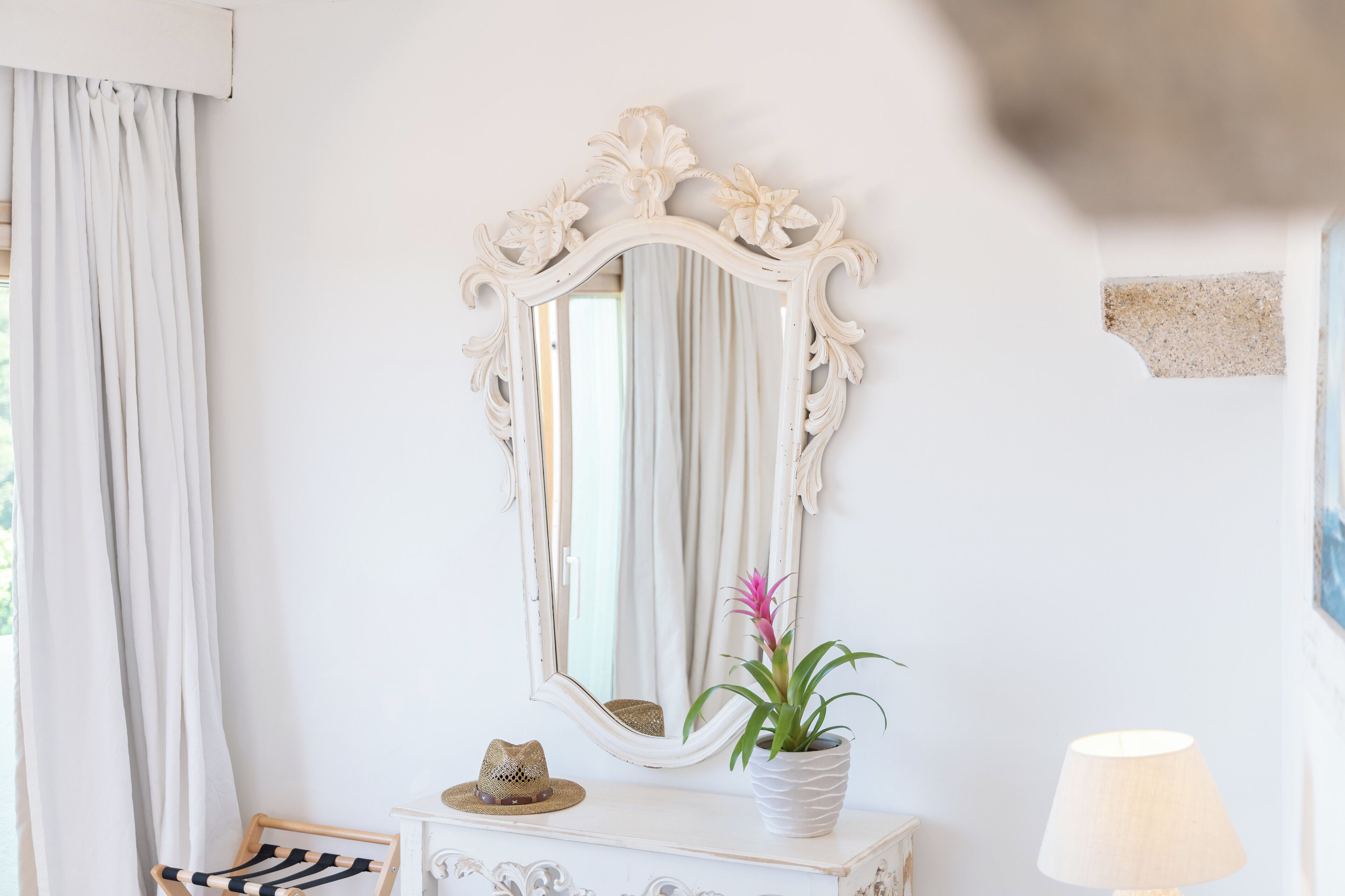 Una habitación con un aparador blanco, un espejo con diseños florales, una planta en maceta, una lámpara y un sombrero sobre el aparador.