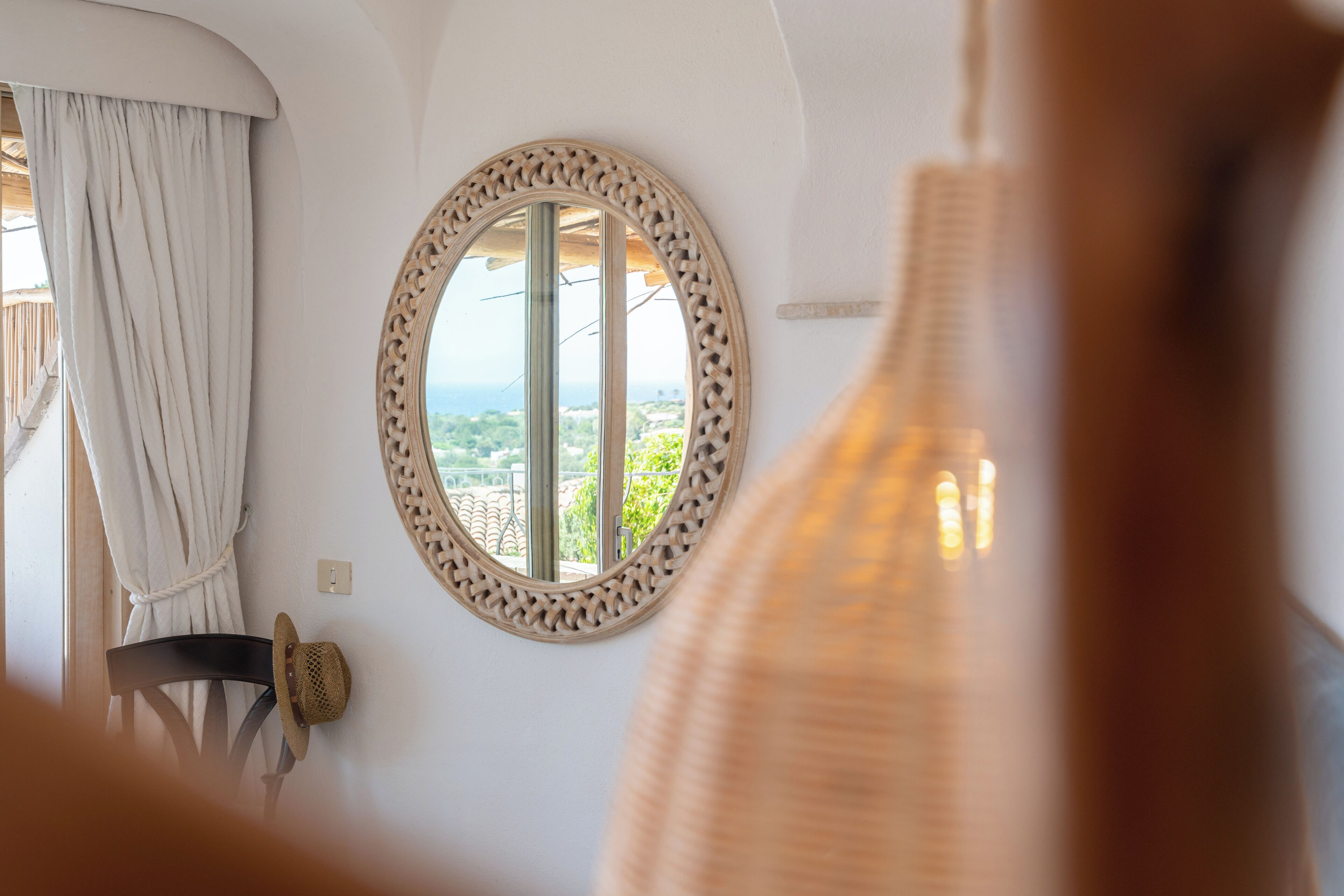 Una habitación con un espejo grande y redondo, vistas por una ventana y una silla de mimbre.