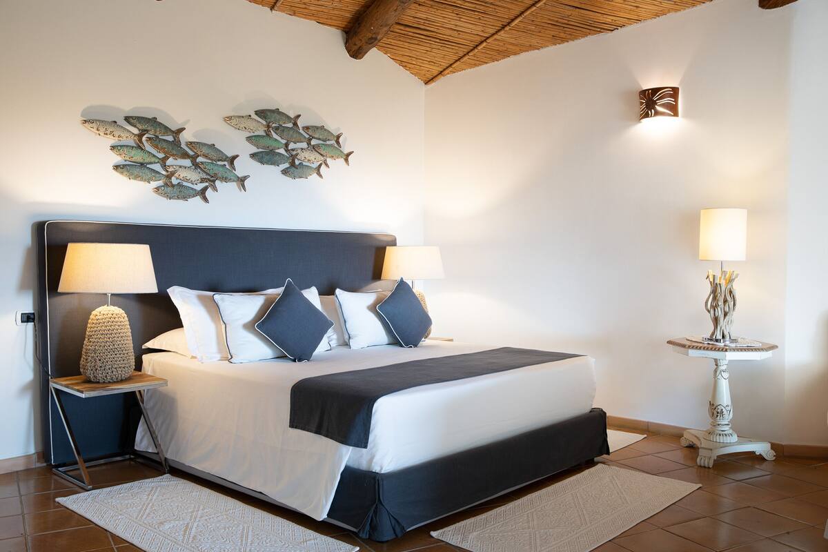 Une chambre à coucher bien rangée, avec un lit, deux lampes de chevet, un élément décoratif mural et une petite table avec une lampe.