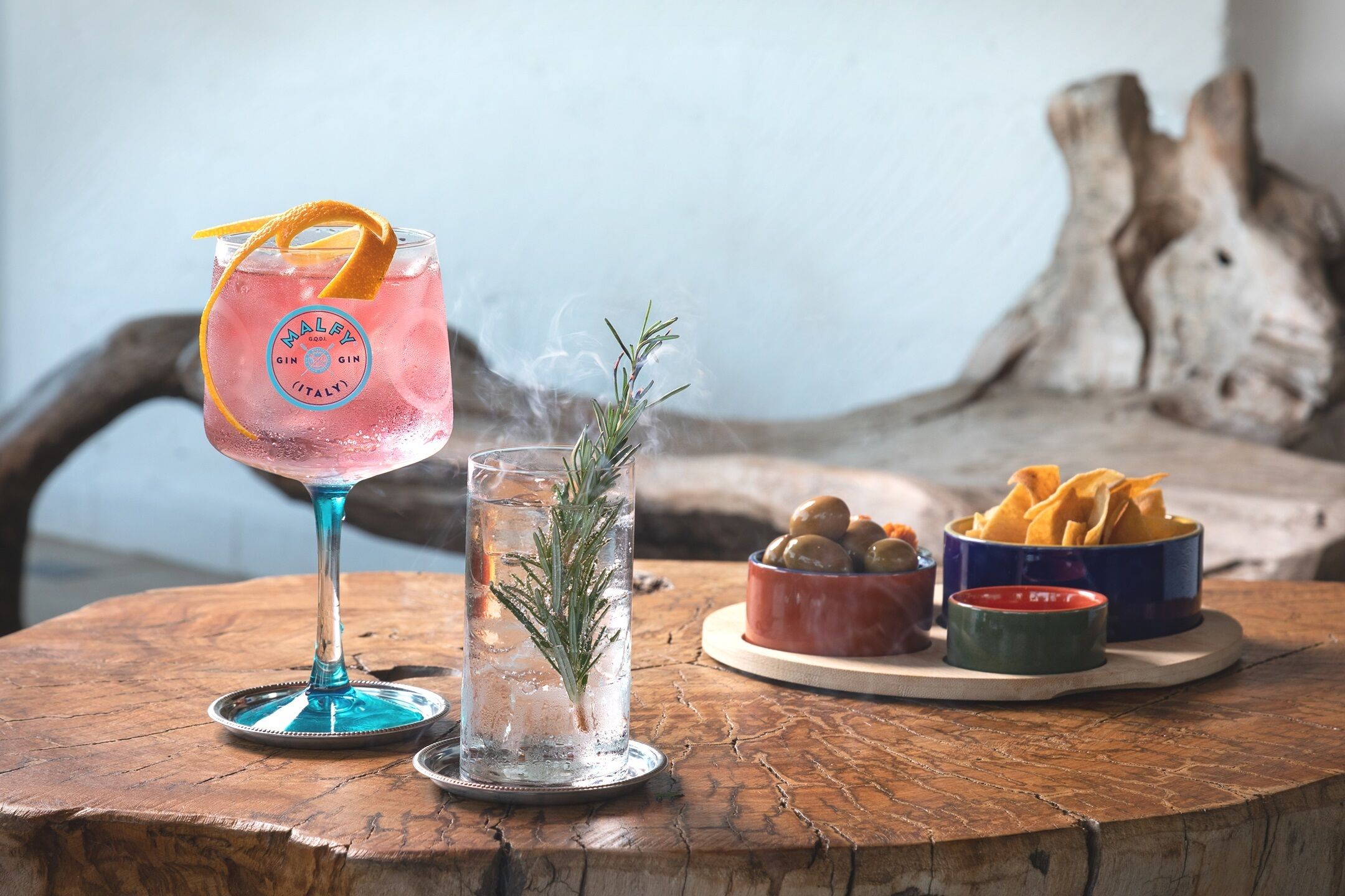 A cocktail glass with a lemon peel garnish, a glass of water with a rosemary sprig, and a tray with chips and olives.