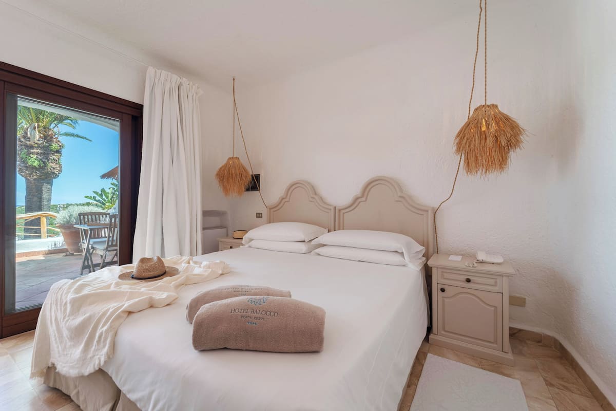 Un lit bien fait, avec une tête de lit blanche, une table de chevet avec une lampe, et une vue sur l’extérieur à travers une porte vitrée.