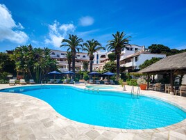 Piscina con sillas de descanso y sombrillas, rodeada de palmeras y edificios.