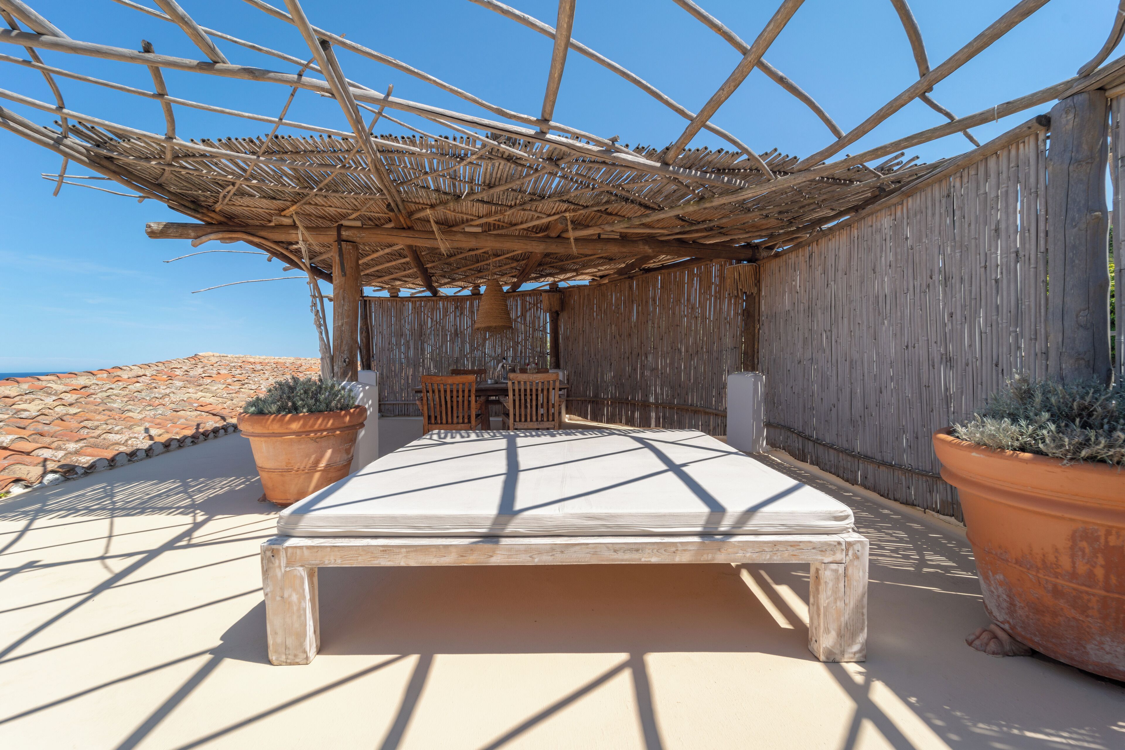 A sunlit outdoor seating area with a wooden bench, two potted plants, and a woven overhead structure.