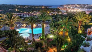 Resort med swimmingpool omgivet af palmer og liggestole, med udsigt over en kystby i skumringen.