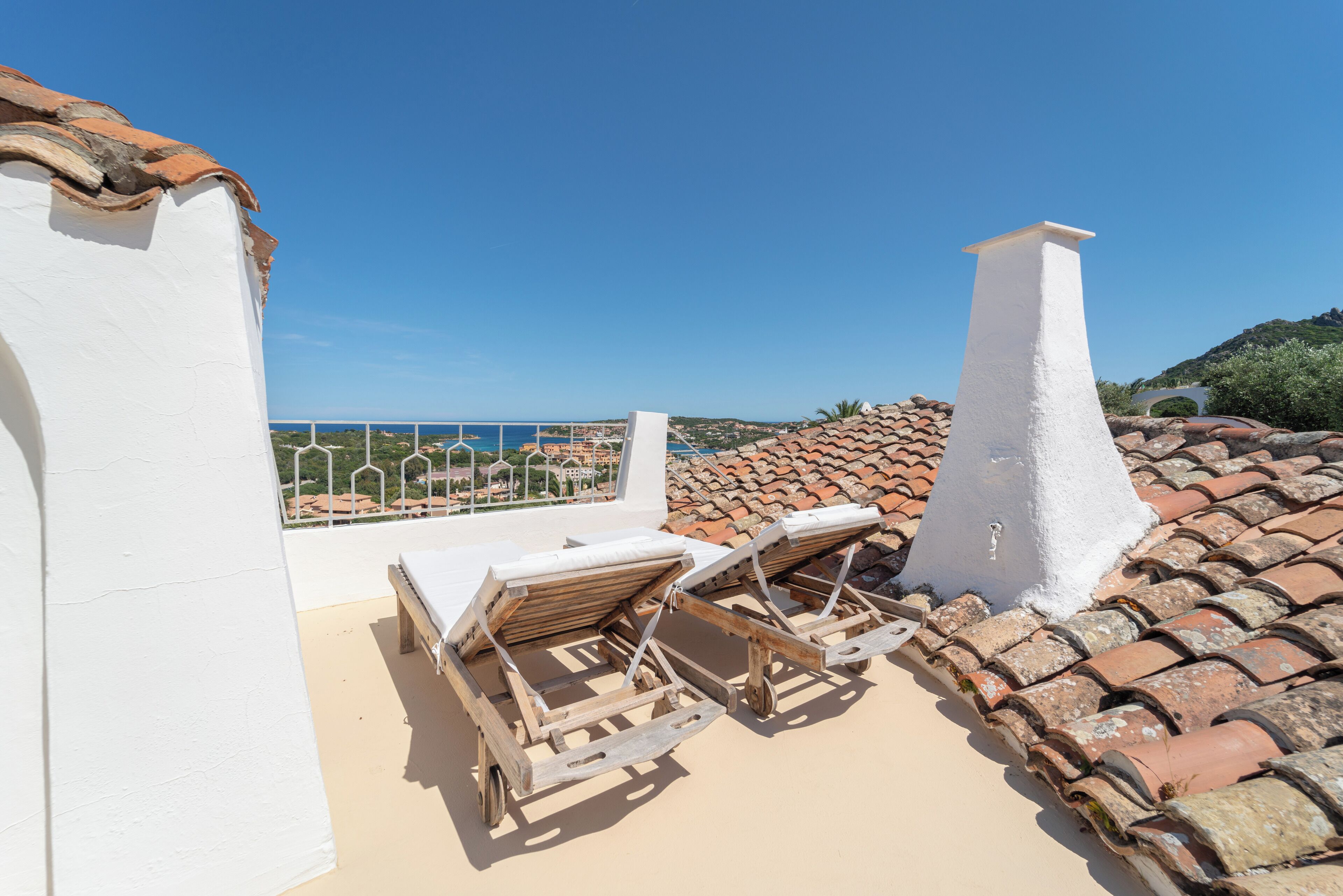 A rooftop terrace with two wooden lounge chairs, a white chimney, and a view of the sea.