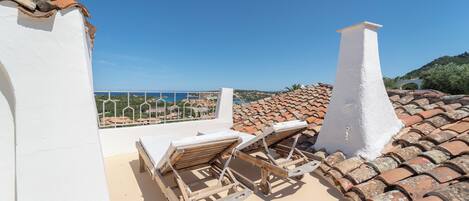 A rooftop terrace with two wooden lounge chairs, a white chimney, and a view of the sea.