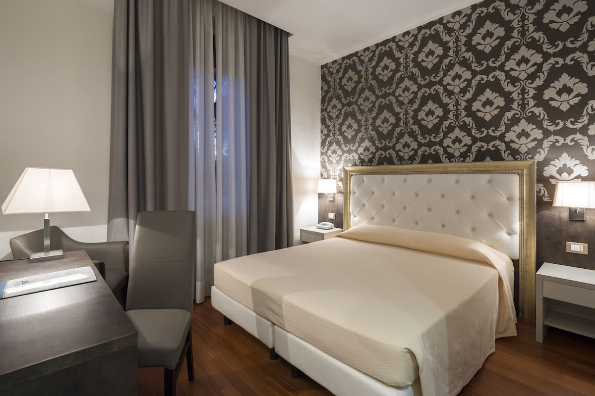 A neatly arranged hotel room with a large bed, a desk, a chair, and a nightstand with lamps.