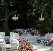 Espaço para casamento ao ar livre, com cadeiras brancas, arranjos florais e lustres.