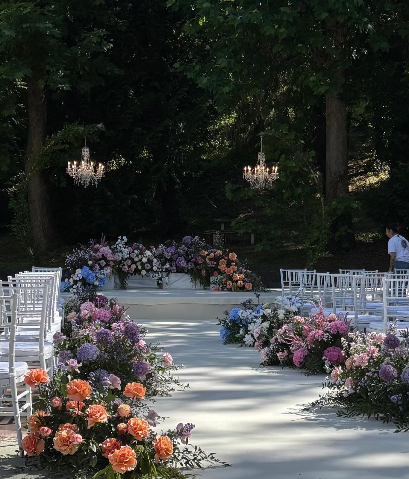 Un pasillo nupcial al aire libre con sillas blancas, arreglos florales y candelabros.
