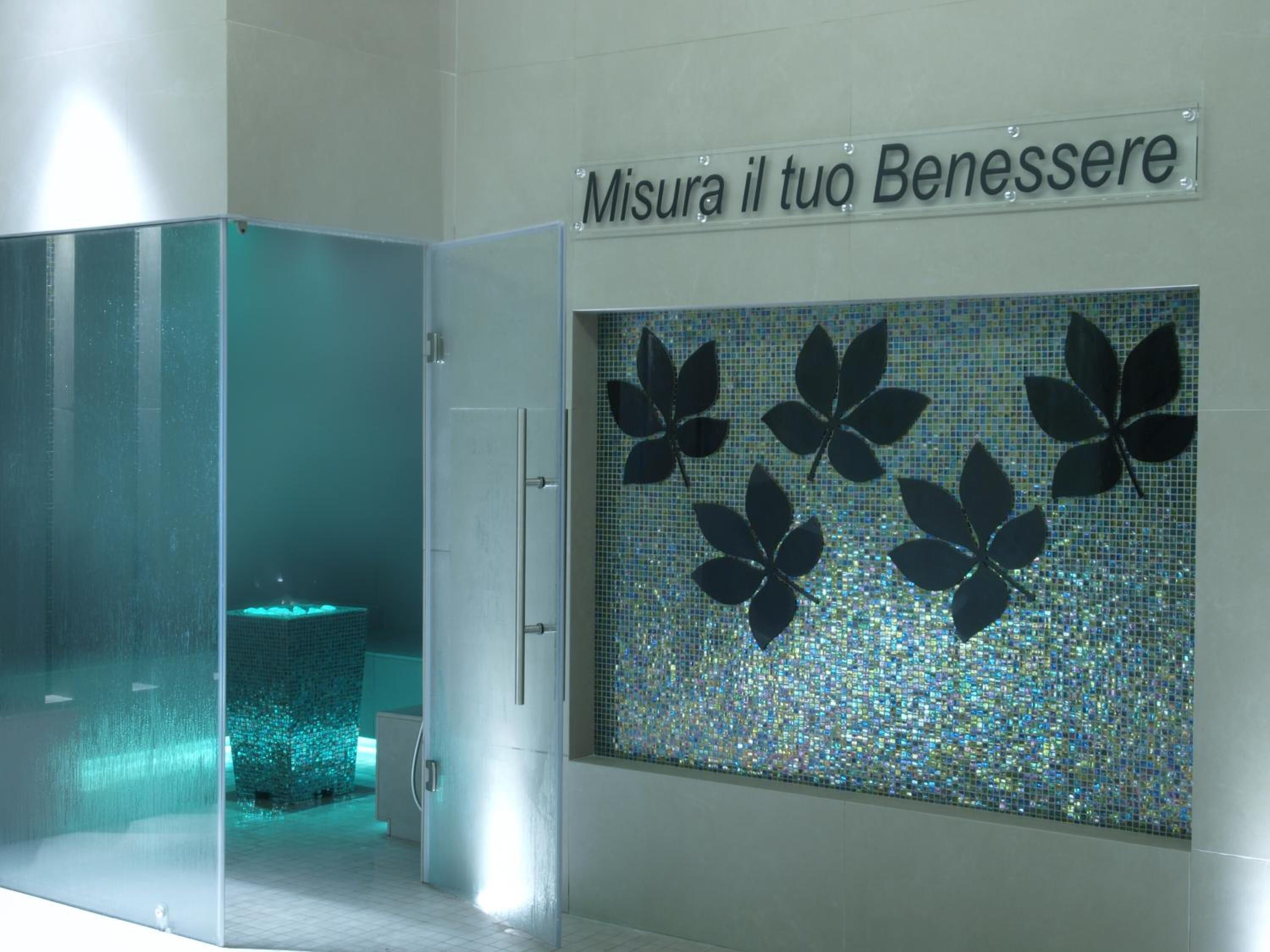 Un baño con puerta de vidrio esmerilado, pared de azulejos con diseño de mosaico y un letrero que dice 'Misura il tuo Benessere'.